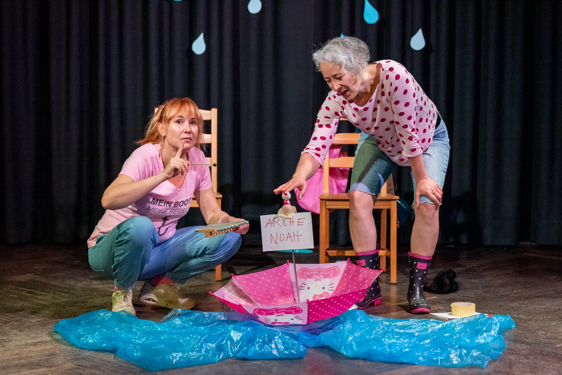 Jana und Jule spielen ARCHA mit einem Regenschirmboot vor einer blauen Regenkulisse